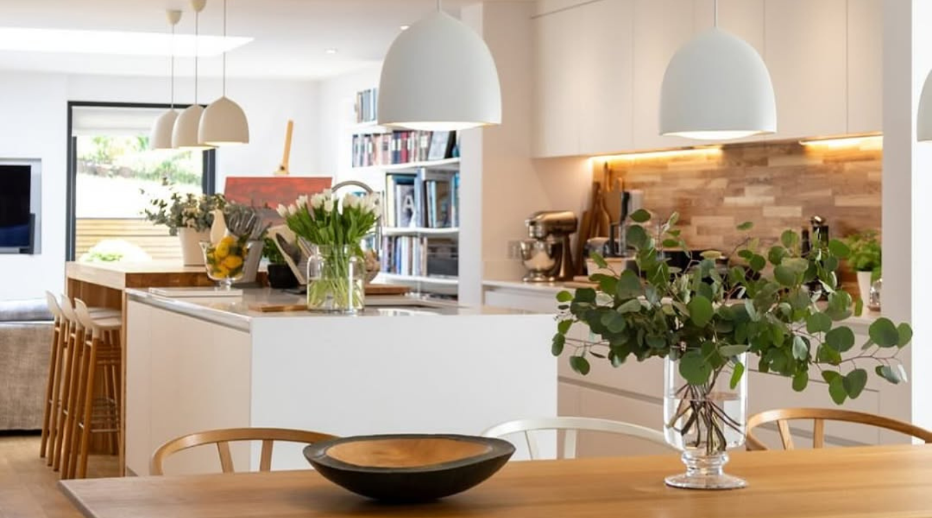 Modern kitchen with white cabinets, wood accents, and pendant lights. A dining table with a bowl and vase is in the foreground.