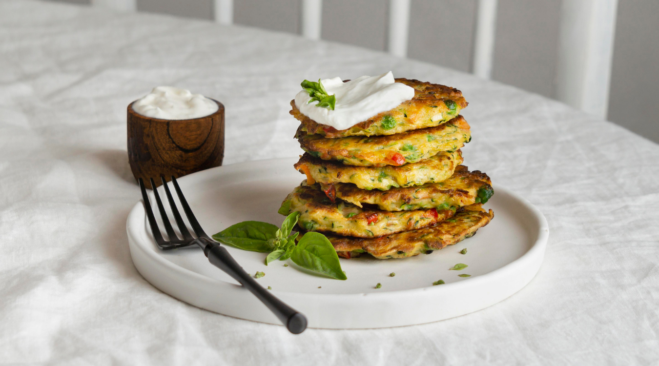 A tall stack of savory pancakes crowned with a dollop of sour cream, presented on a white plate alongside a small dish of dipping sauce and a fork ready to serve. Stack of savory pancakes topped with sour cream, served on a white plate with a dipping sauce and a fork.