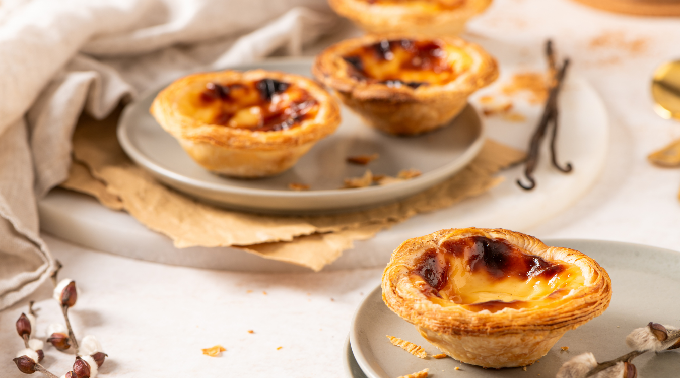 Golden-brown Portuguese custard tarts arranged on plates, garnished with fragrant vanilla bean and delicate sprigs of greenery, set against a soft, light-colored surface. Golden-brown Portuguese custard tarts on plates, with vanilla bean, and sprigs of plants on a light surface.