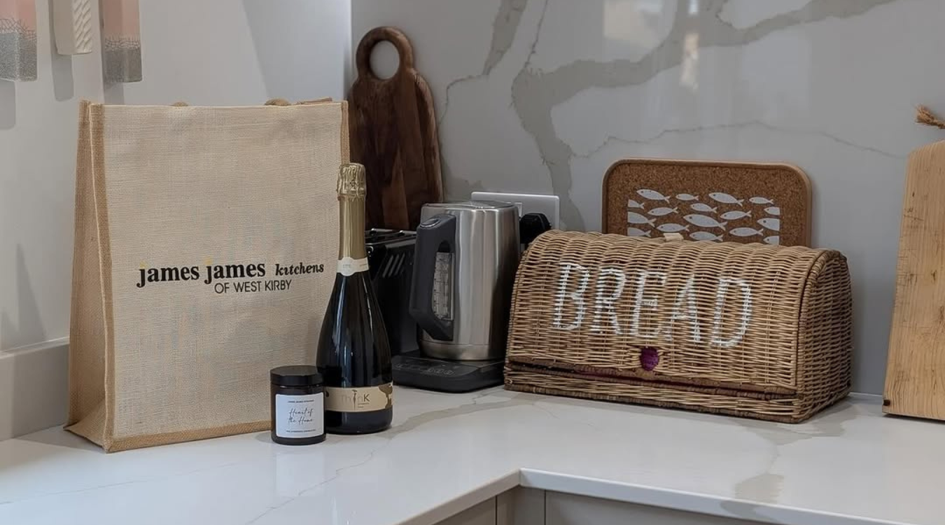 Kitchen countertop with food items: a paper bag, champagne, jam jar, kettle, bread basket, and cutting boards.