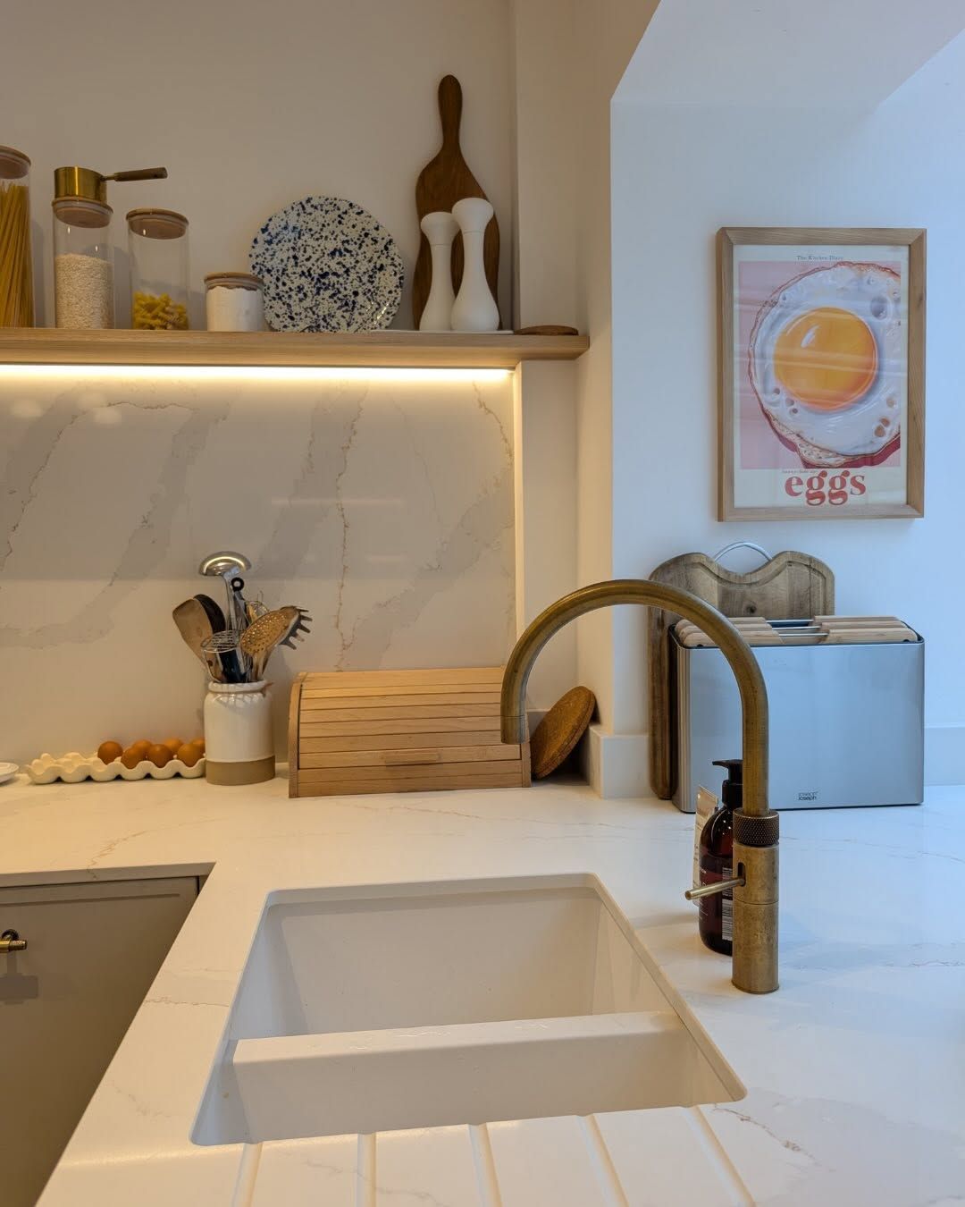 A bright kitchen features a white sink with a brass faucet, marble countertops, and a shelf with jars and decor.