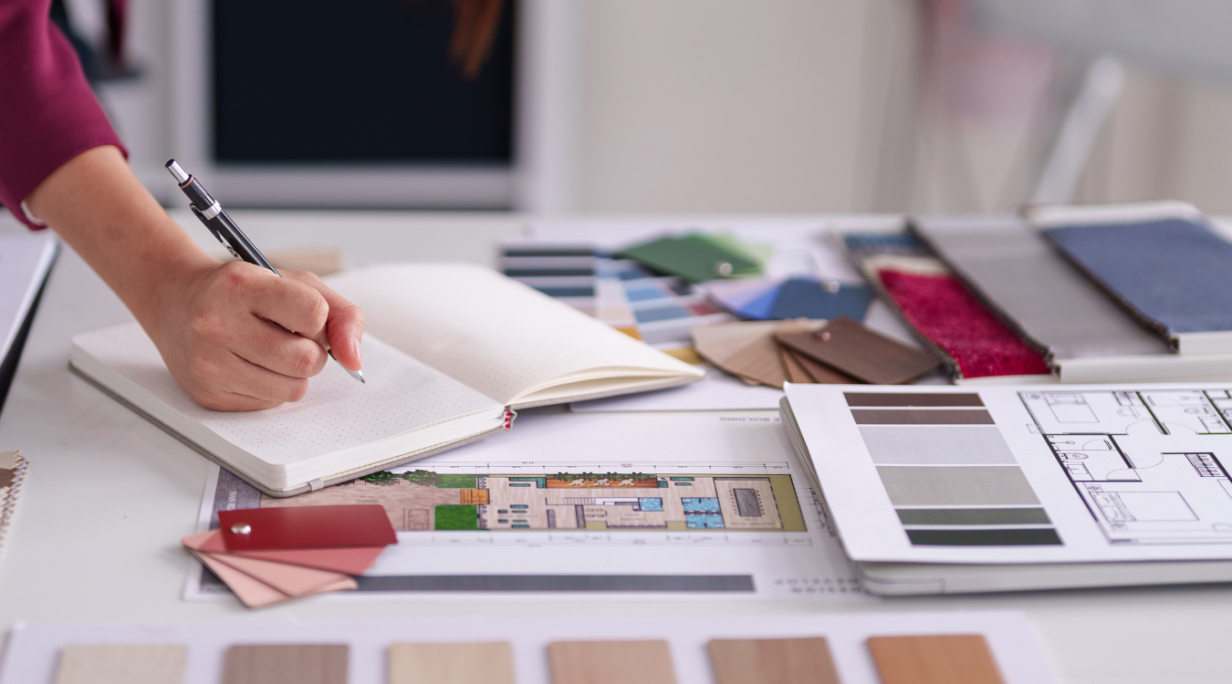Person writing in a notebook on a desk with color swatches, blueprints, and a computer monitor.