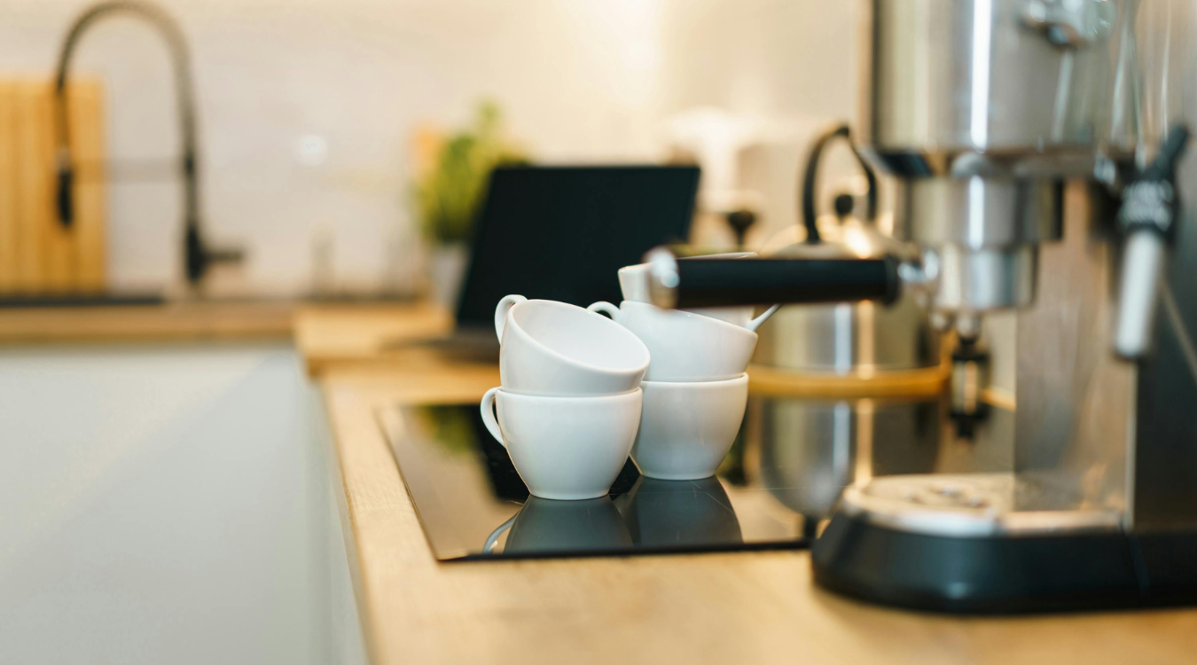 White ceramic cups stacked on a black stovetop next to a stainless steel espresso machine in a modern kitchen.