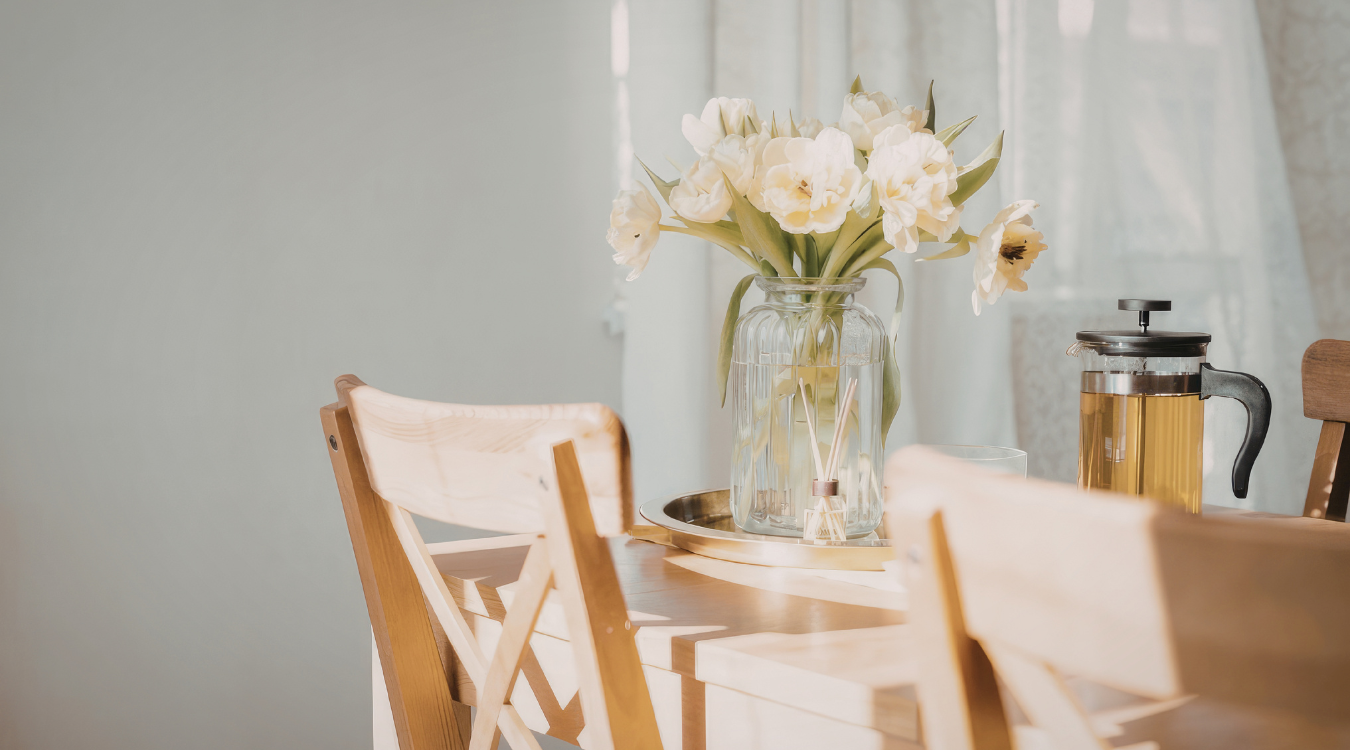 A dining table styled with a vase of white flowers, a French press, and wooden chairs positioned beside a light-filled window. Dining table with vase of white flowers, french press, and wooden chairs by a window.