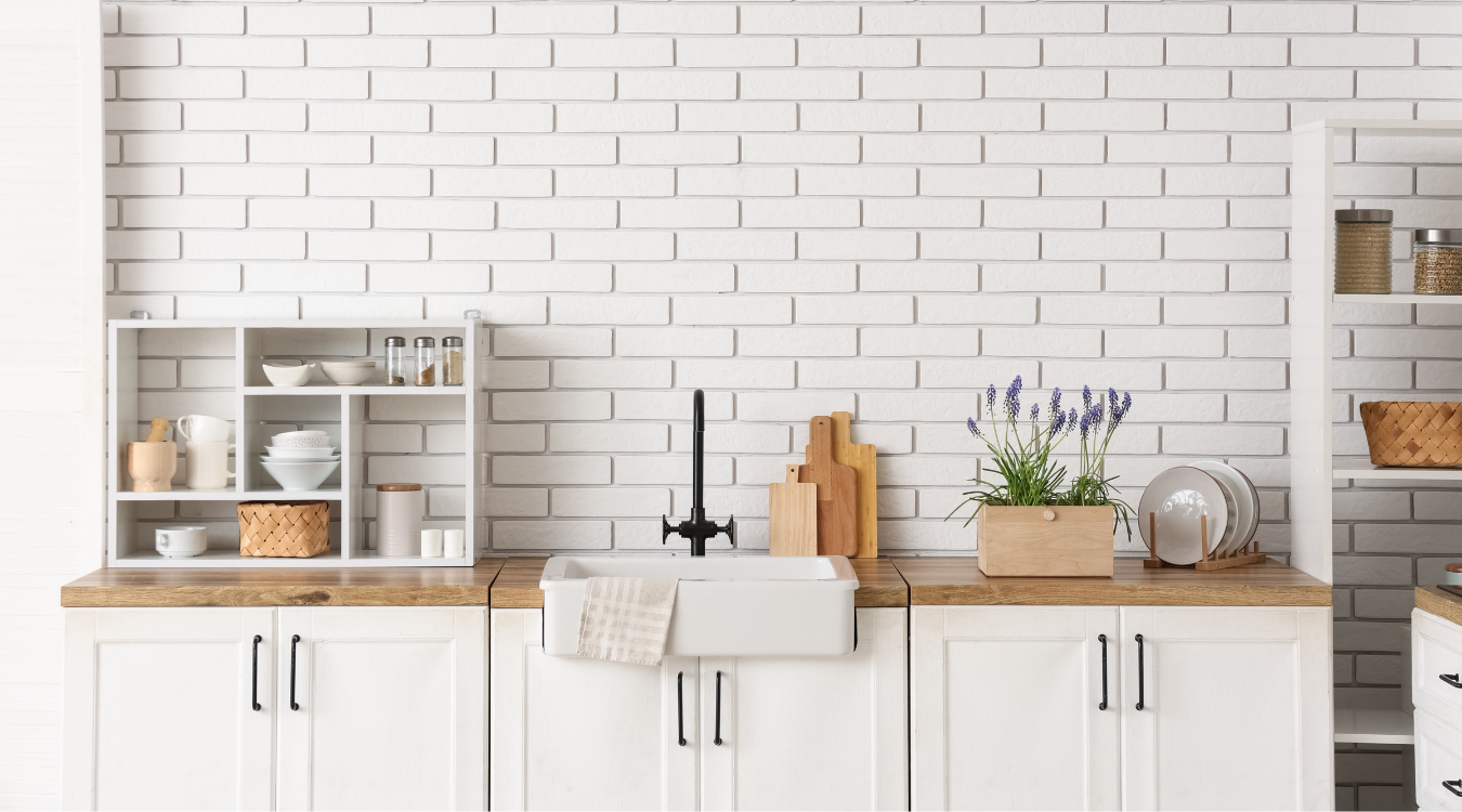 A white kitchen set against an exposed brick wall, featuring a farmhouse sink, wooden worktops, crisp white cabinetry, and open shelves styled with décor. White kitchen with brick wall, farmhouse sink, wood countertops, white cabinets, and shelves with décor.