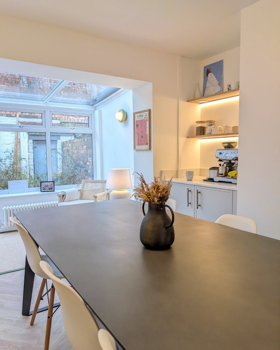 A dining area with a dark table, white chairs, a black vase, and a lit coffee station against a conservatory wall.