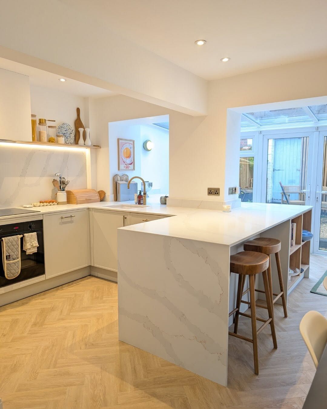 A modern kitchen with a large white marble-topped island, wooden bar stools, and herringbone flooring.