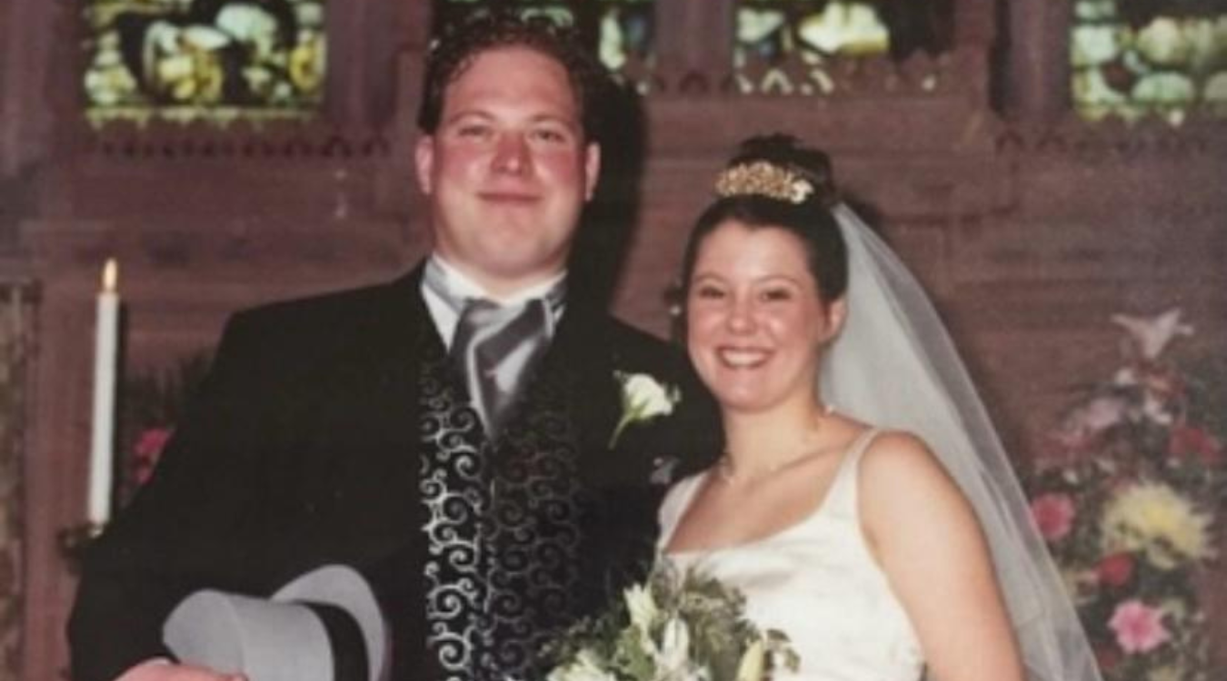 A bride and groom pose for a portrait in a church, smiling in wedding attire with a bouquet and top hat.