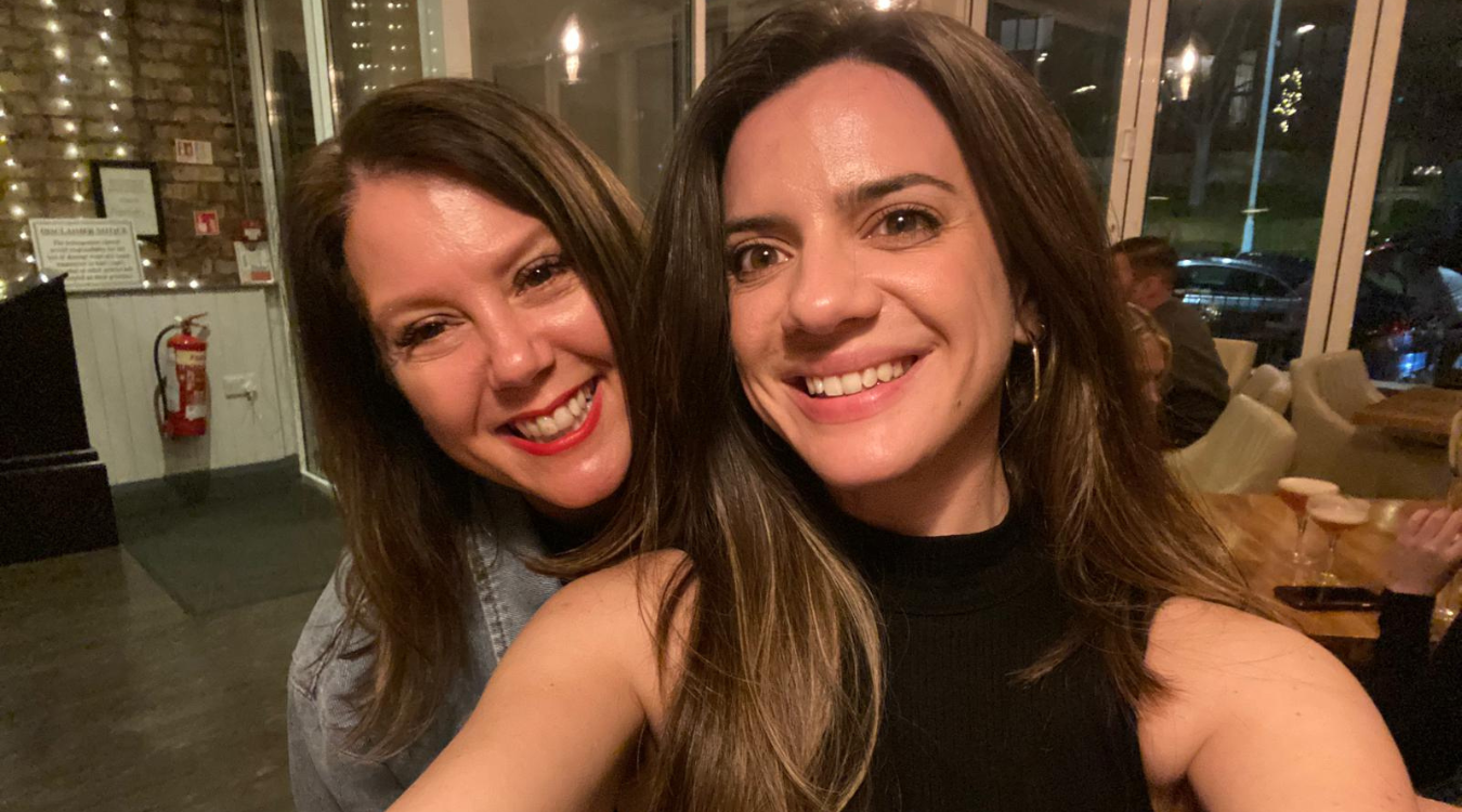 Two smiling people pose for a selfie in an indoor setting with string lights and furniture in the background.