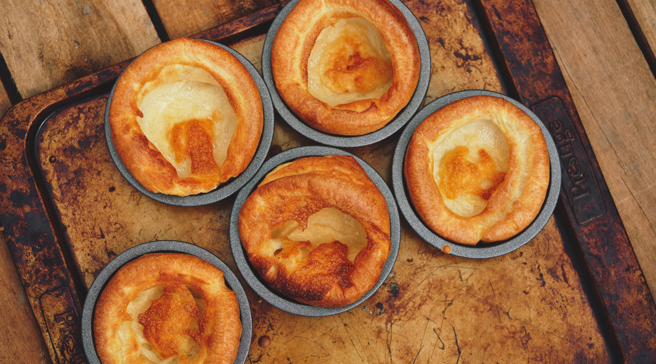 Golden, puffed Yorkshire puddings baked in individual tins, resting on a rustic wooden surface with their crisp, beautifully browned tops on display. Yorkshire puddings in individual tins on a rustic, wooden surface. Golden brown, puffed-up pastry.