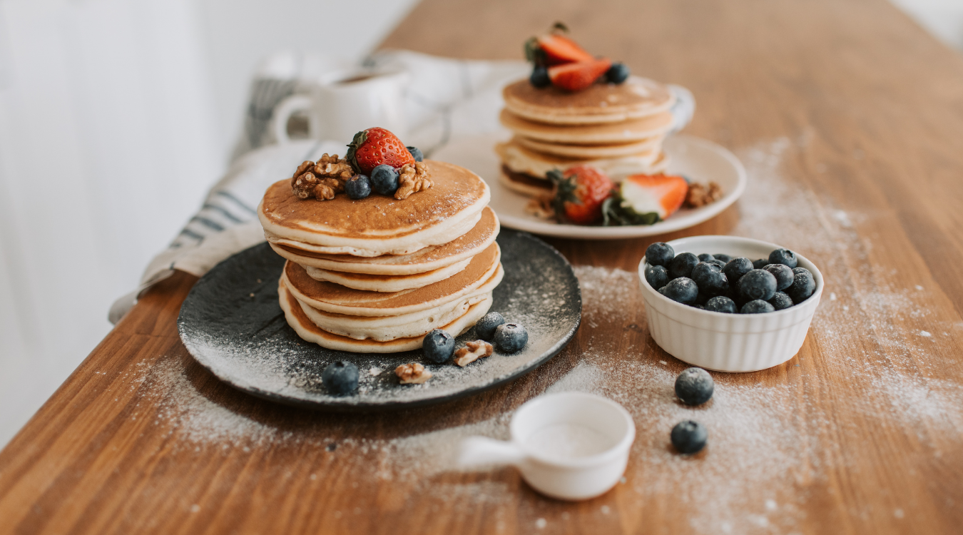 A stack of fluffy pancakes topped with fresh blueberries, sliced strawberries, and crunchy walnuts sits on a rustic wooden table, accompanied by a steaming cup of coffee and a small bowl of blueberries on the side. Pancakes stacked with blueberries, strawberries, and walnuts on a wooden table, with a cup of coffee and blueberries nearby.