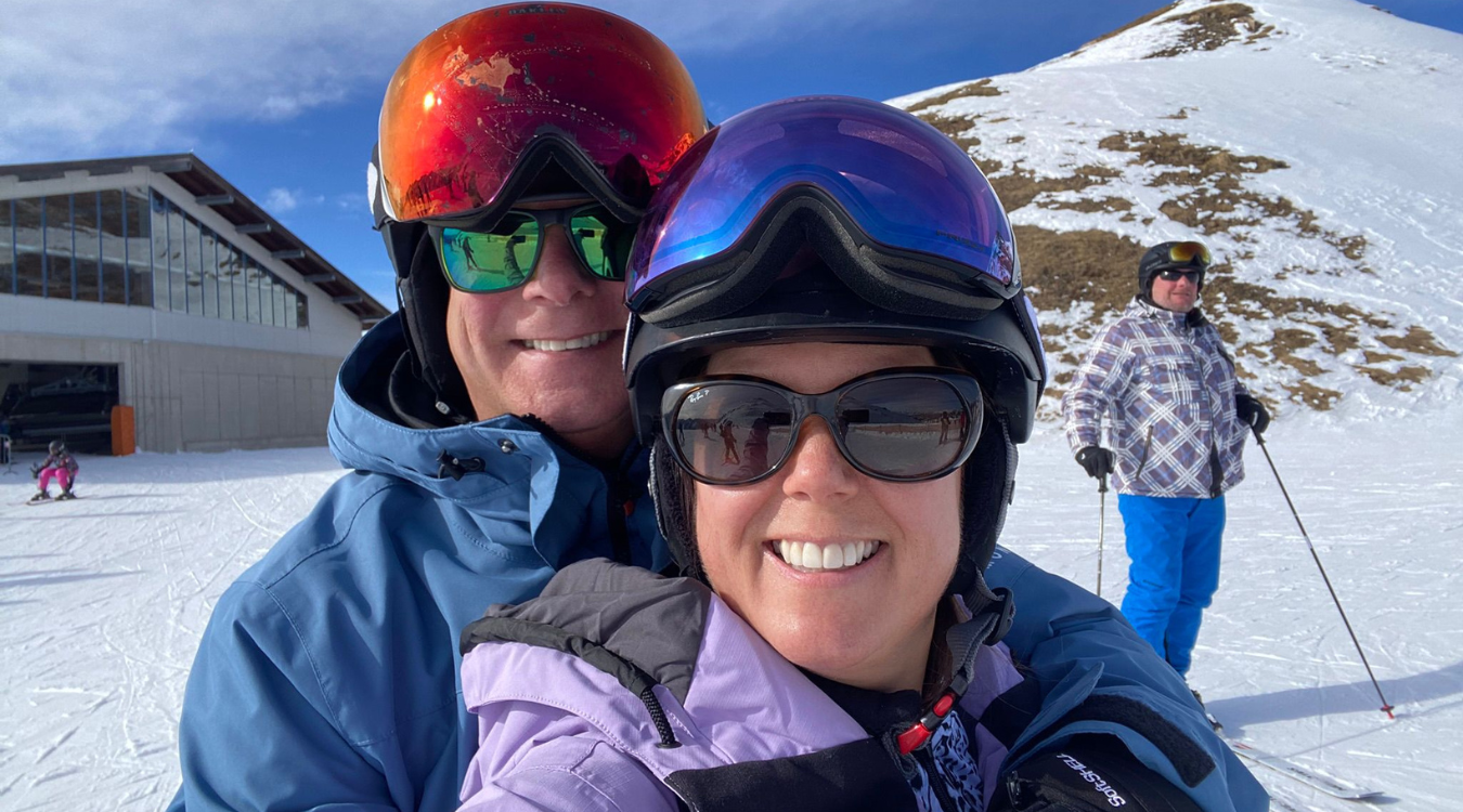 A couple in ski gear and goggles pose for a selfie at a snowy ski resort, with another skier in the background.
