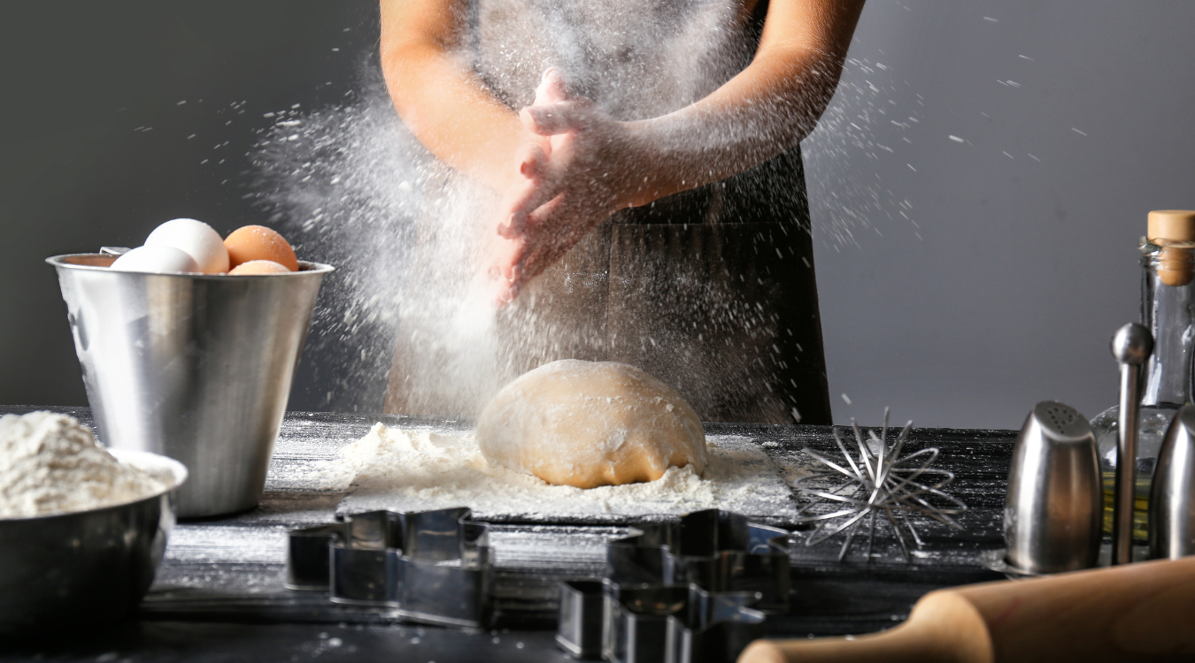 Person clapping hands, dusting flour over a dough on a table with baking ingredients and tools.