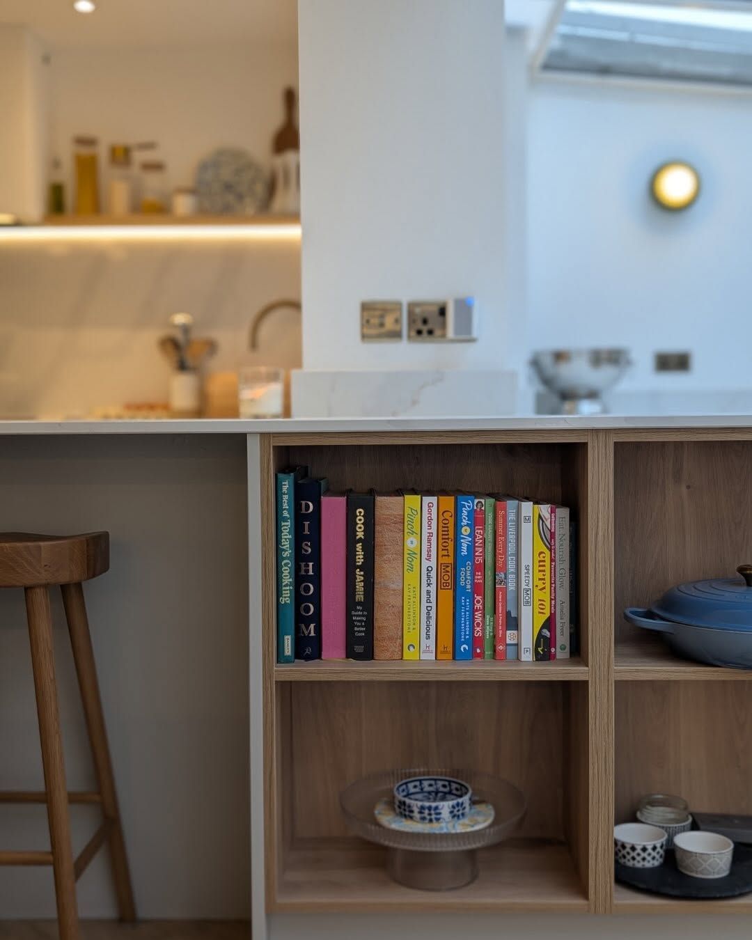 A kitchen island with a wooden bookshelf displaying cookbooks, a bowl, and ceramic dishes, next to a bar stool.