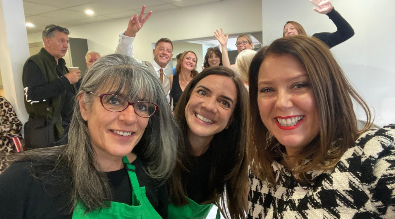 A group of smiling people pose for a selfie in a brightly lit room; some wear green aprons, and others wave in the back.