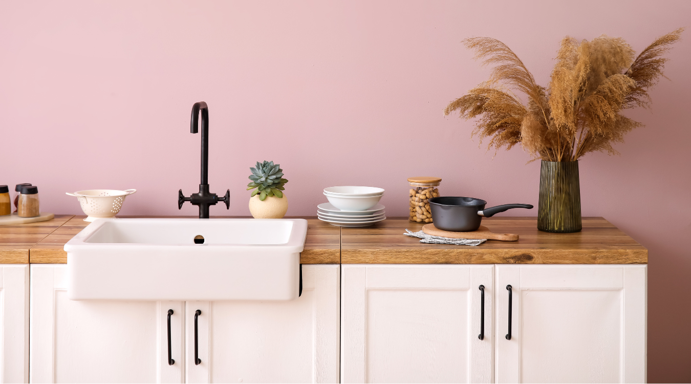 White kitchen sink and cabinets with wooden countertop; pink wall; decor includes plants and a pot.
