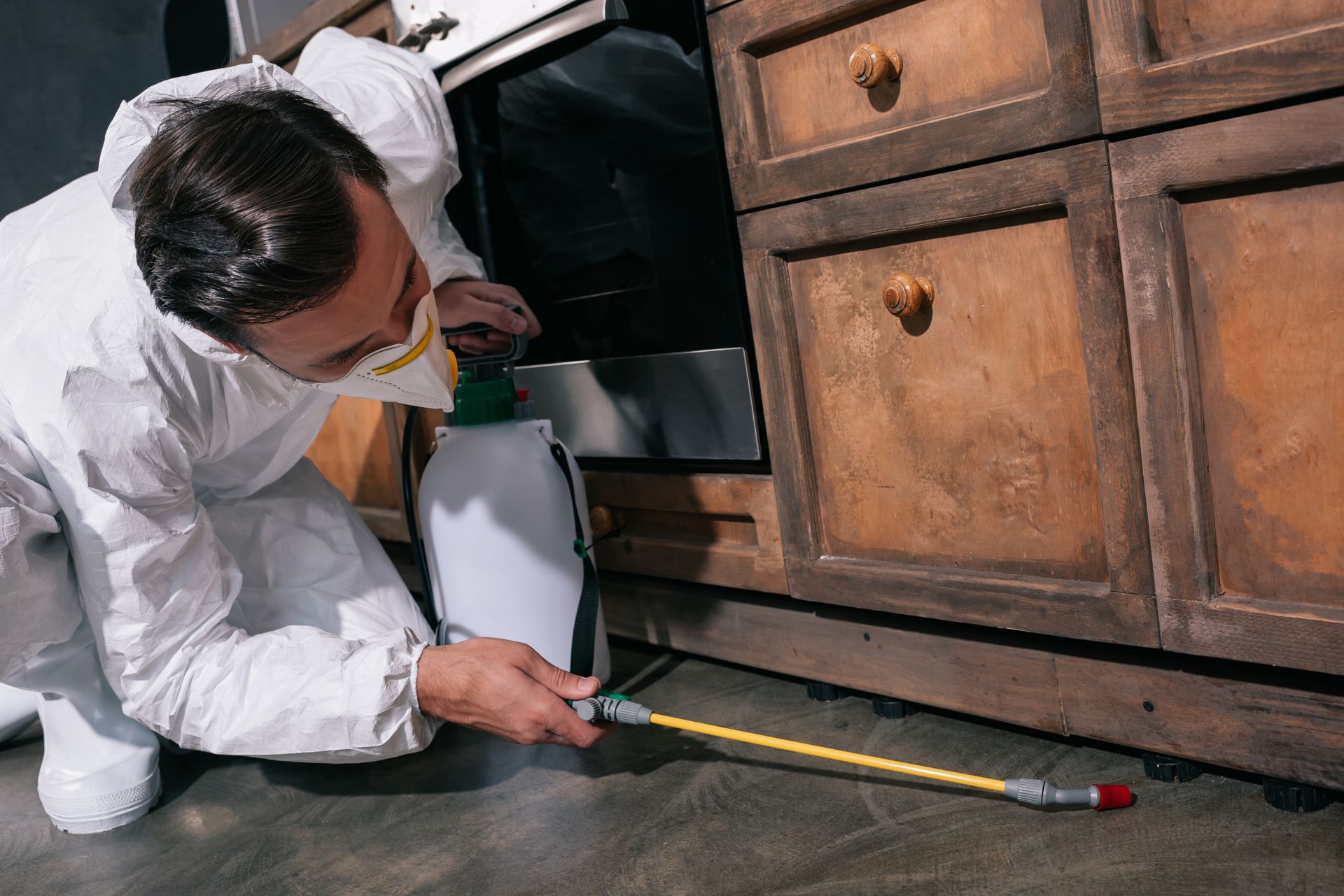 Pest control worker in uniform spraying pesticides under cabinet in kitchen
