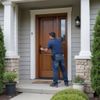 Man opening a wooden door on a house porch. He's wearing a blue shirt and jeans. Gray siding, stone pillars.