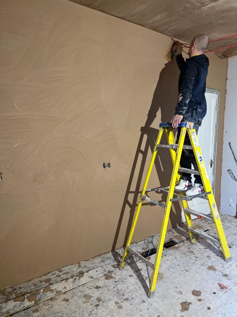 Person plastering a wall while standing on a yellow ladder in a room under construction.