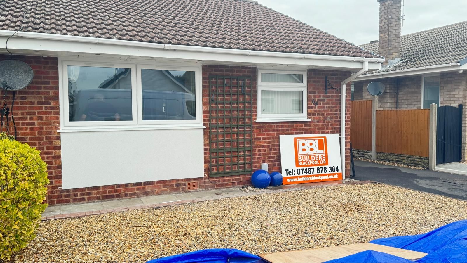 Brick bungalow with white trim, windows, and a for-sale sign on a gravel yard.