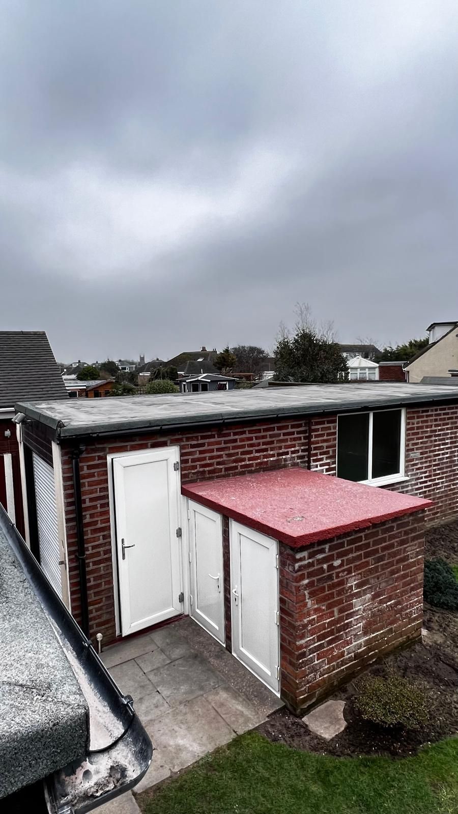 Brick building with white doors, window, and red flat roof under a cloudy sky.