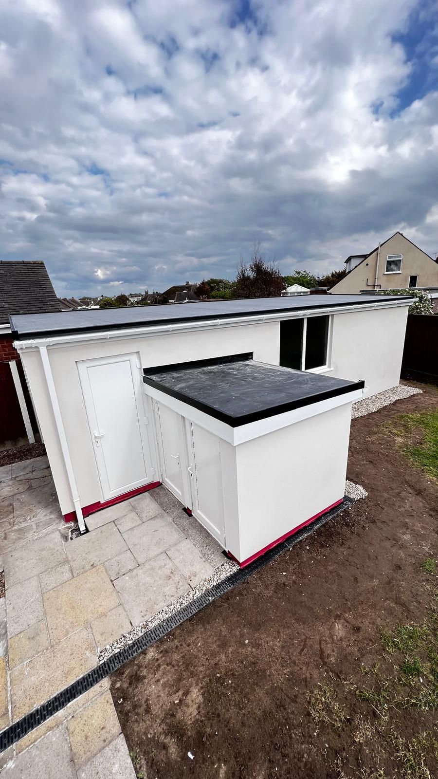 White shed with a black flat roof and small canopy extension under a cloudy sky.