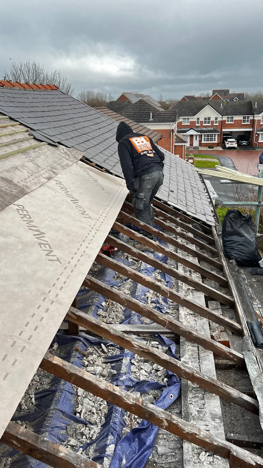 A person repairs a roof, installing underlayment on exposed rafters. Overcast day.