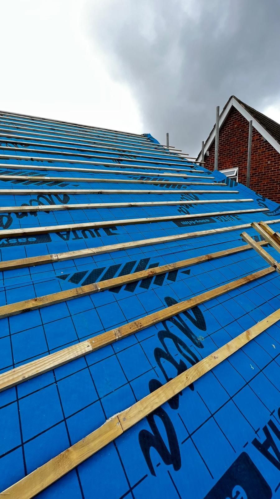Roof under construction; blue underlayment and wooden battens on sloped roof, cloudy sky.