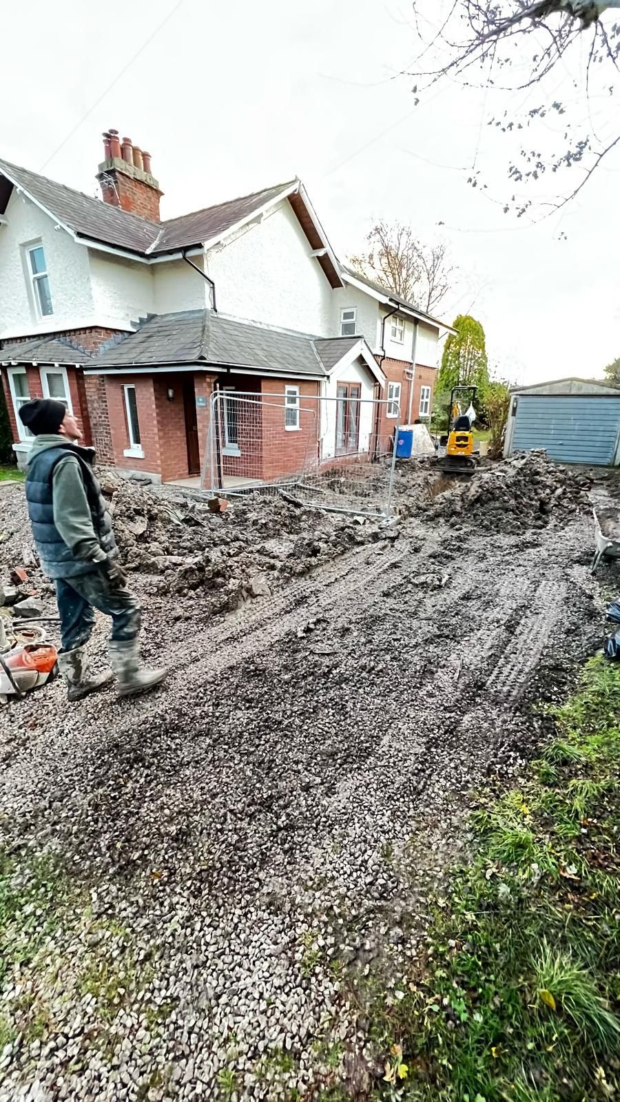 Man watches construction site. Brown dirt, brick building, yellow excavator, cloudy sky.