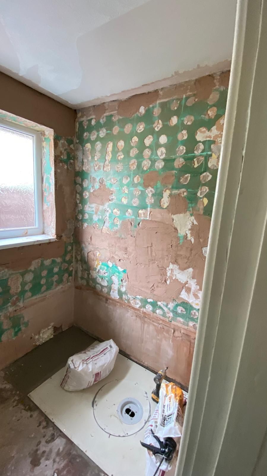 Bathroom under renovation; walls with exposed green patches, cement floor, window, and supplies.