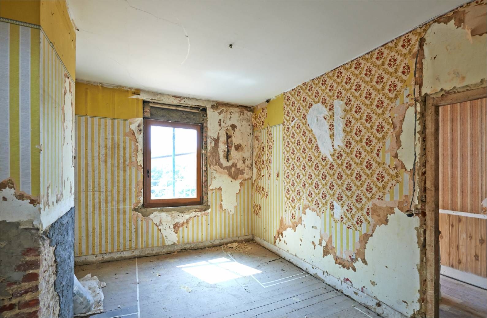 Room with peeling wallpaper, window, and doorway, appearing under renovation.