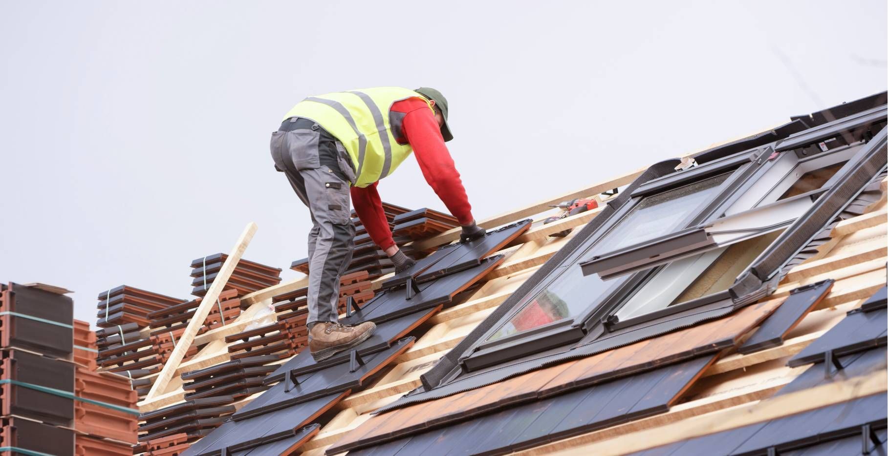 Roofer in safety vest laying tiles on a rooftop with skylights, against a cloudy sky.