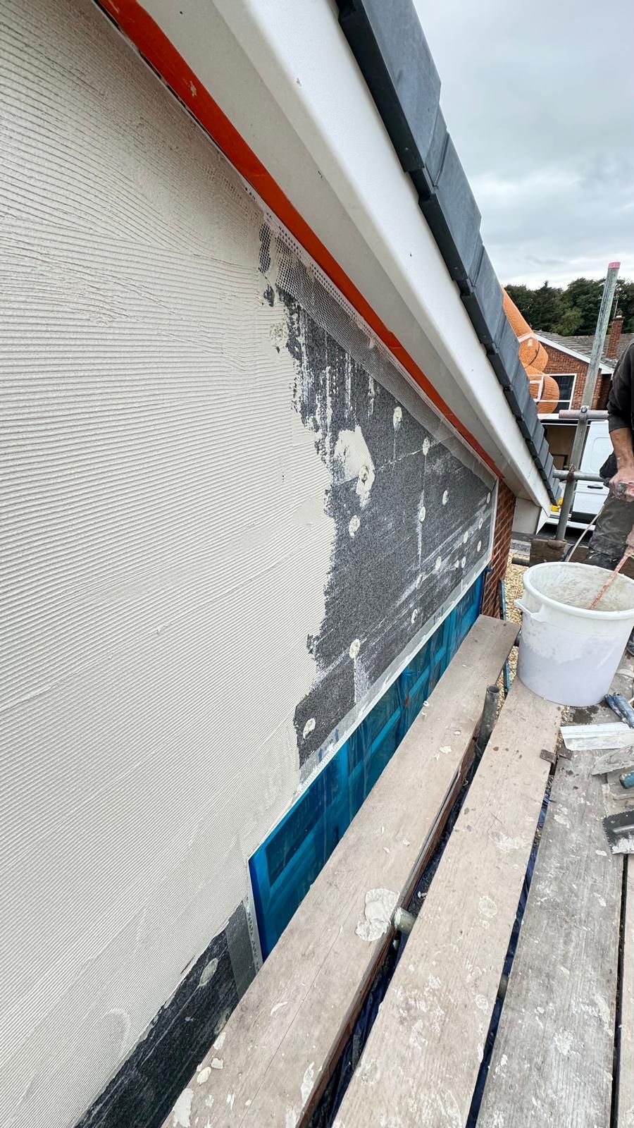 Exterior wall being plastered, blue trim visible, person on scaffolding.