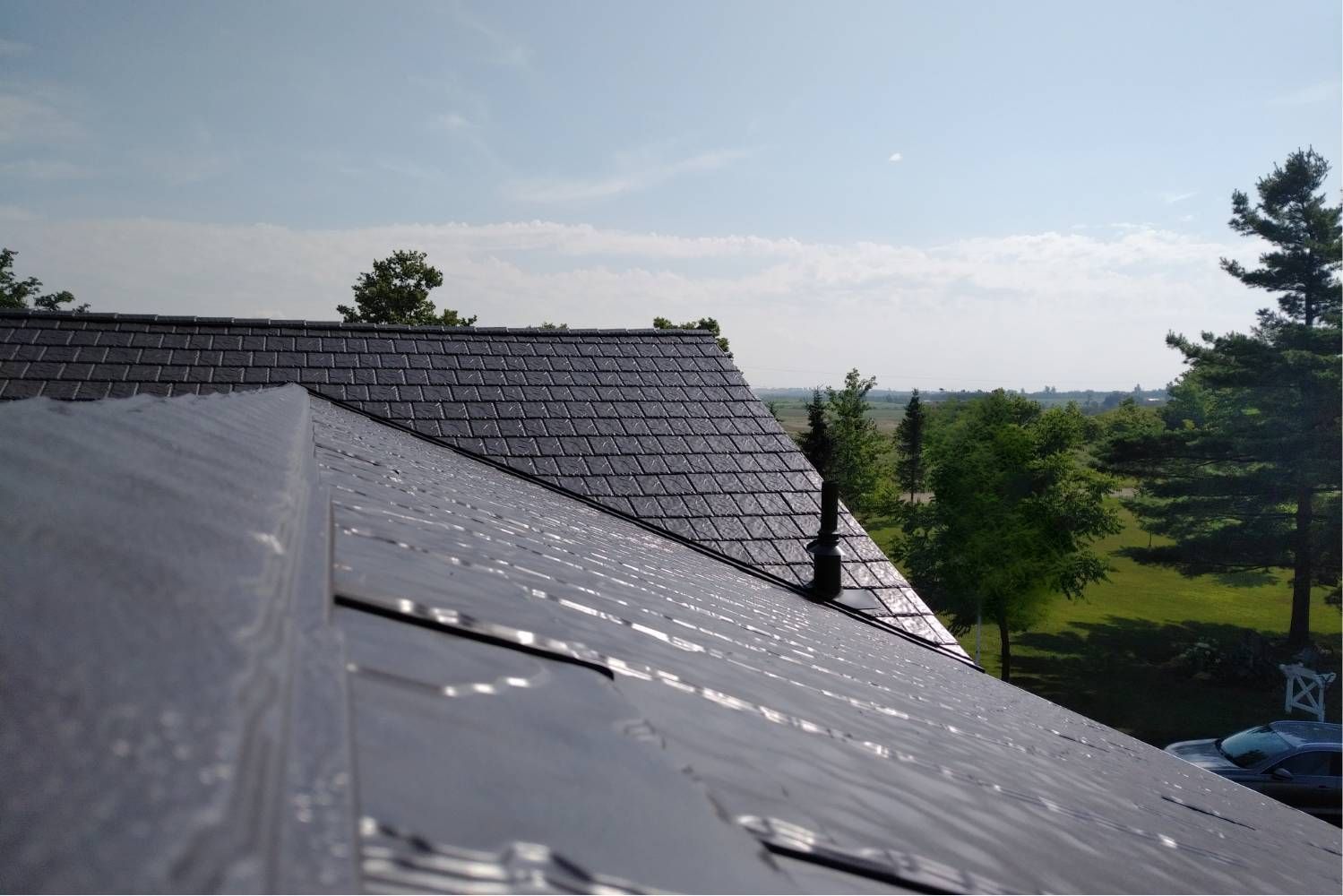 Dark roof with light reflections against a blue sky, trees in background.