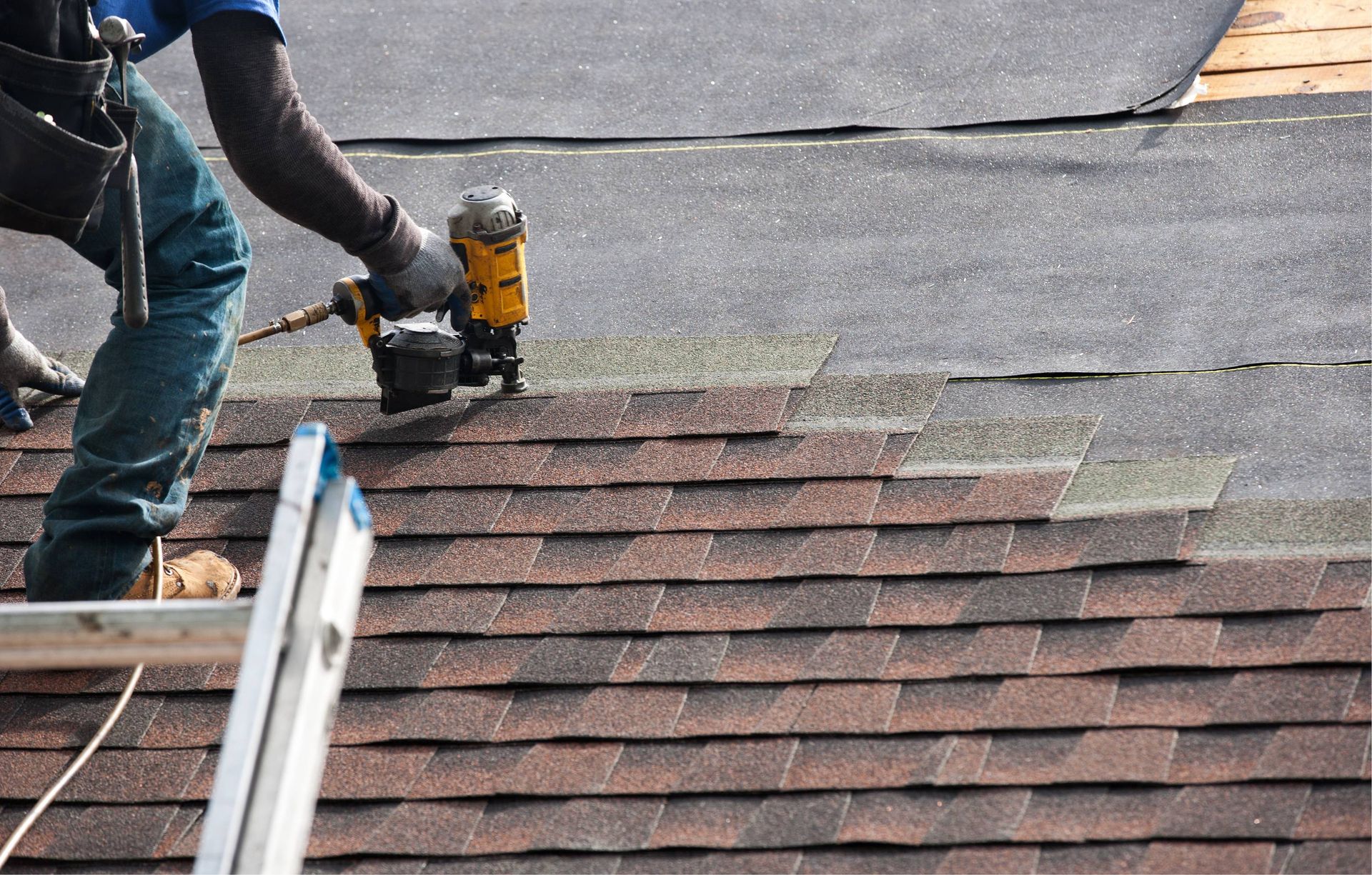 Roofer using a nail gun to install brown asphalt shingles on a roof.