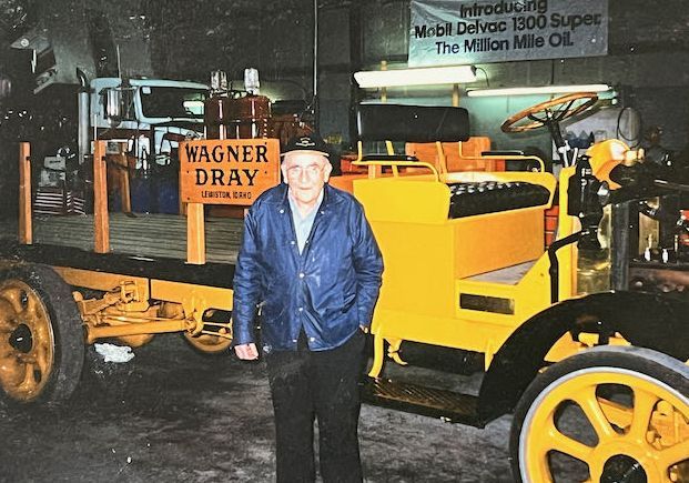 A person stands in front of a restored yellow vintage flatbed truck labeled