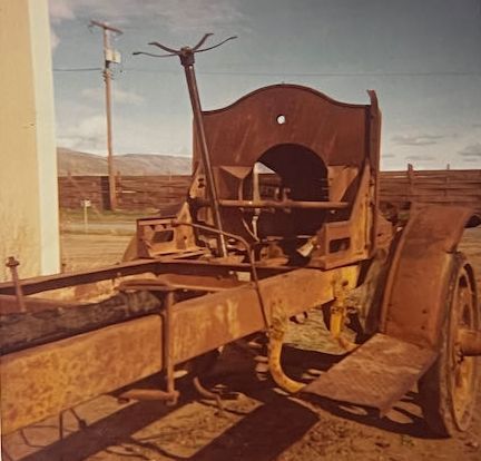 The rusted, stripped chassis of an antique truck sits outdoors on a dirt lot, featuring a tall steering column and wheels.