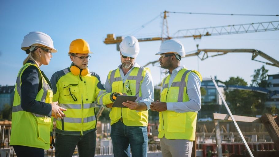 Four construction workers in safety gear discussing a digital tablet.
