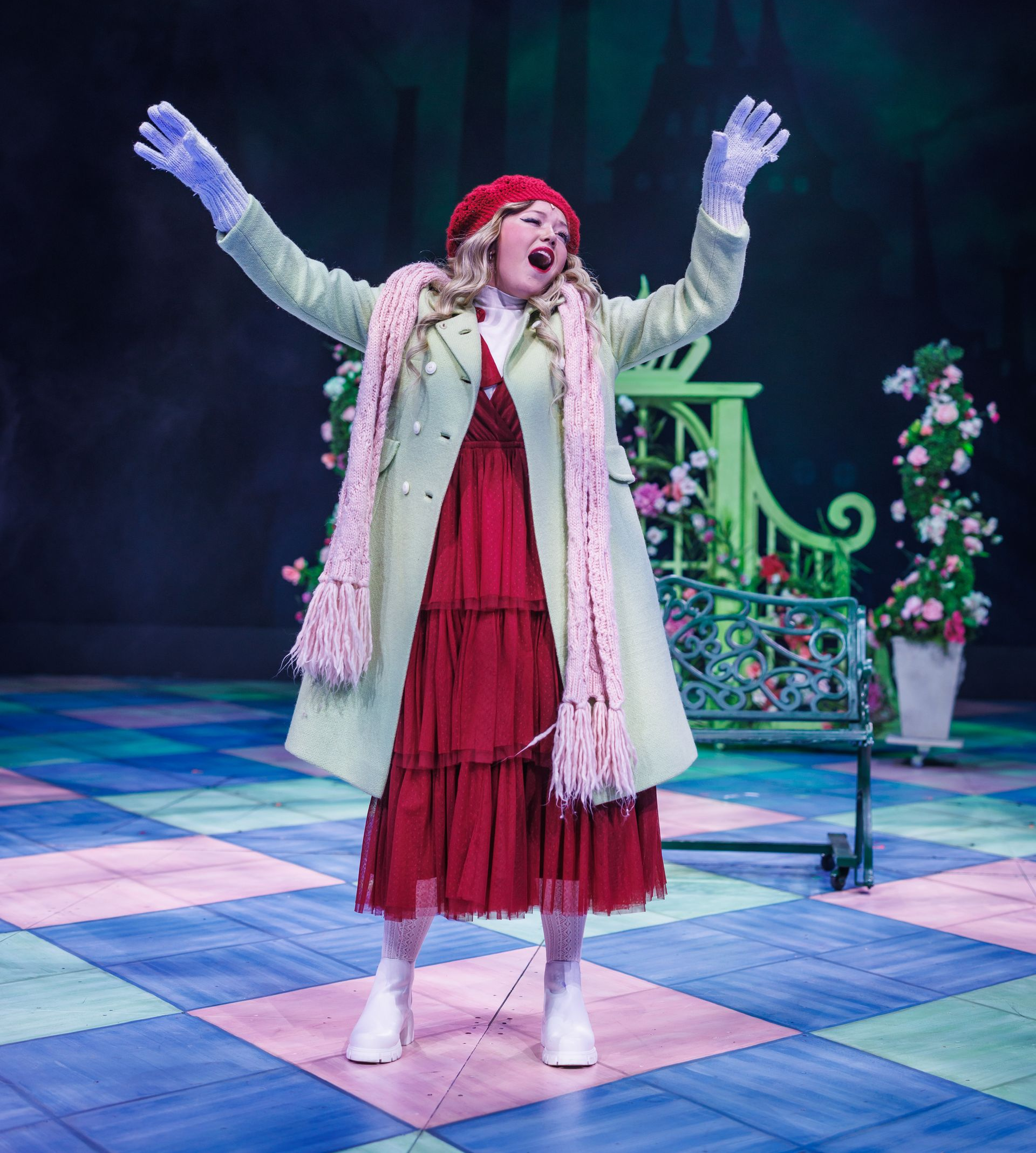 A person in a red dress, light green coat, and beret singing on stage with arms raised, standing on a checkered floor.