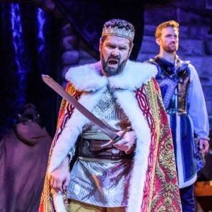 A person dressed as a king in a red and gold robe with a fur collar holds a sword, standing before a person in armor.