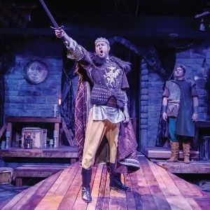 A costumed actor holds a sword aloft on a wooden stage, while a second actor stands in the background on a stone set.