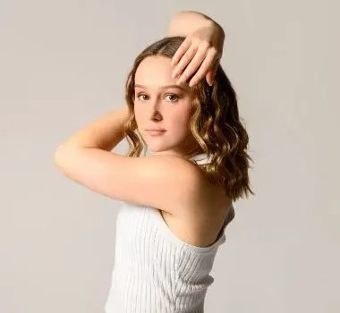 A young person with shoulder-length wavy hair wearing a white ribbed tank top, posing with one hand behind their head.