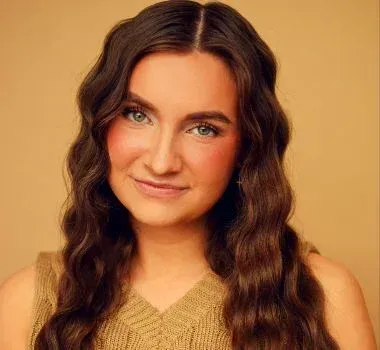 A person with long, wavy brown hair and a subtle smile against a solid tan background, wearing a textured beige top.