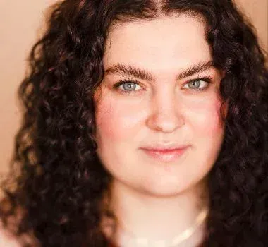 A headshot of a person with dark, curly hair, light eyes, and a neutral expression, posed against a soft beige background.