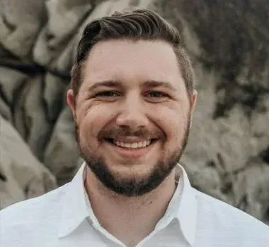 A man with a short beard and styled brown hair smiles while wearing a white collared shirt in front of a rocky background.