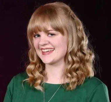 A smiling person with blonde, wavy, shoulder-length hair and bangs, wearing a green top against a black background.