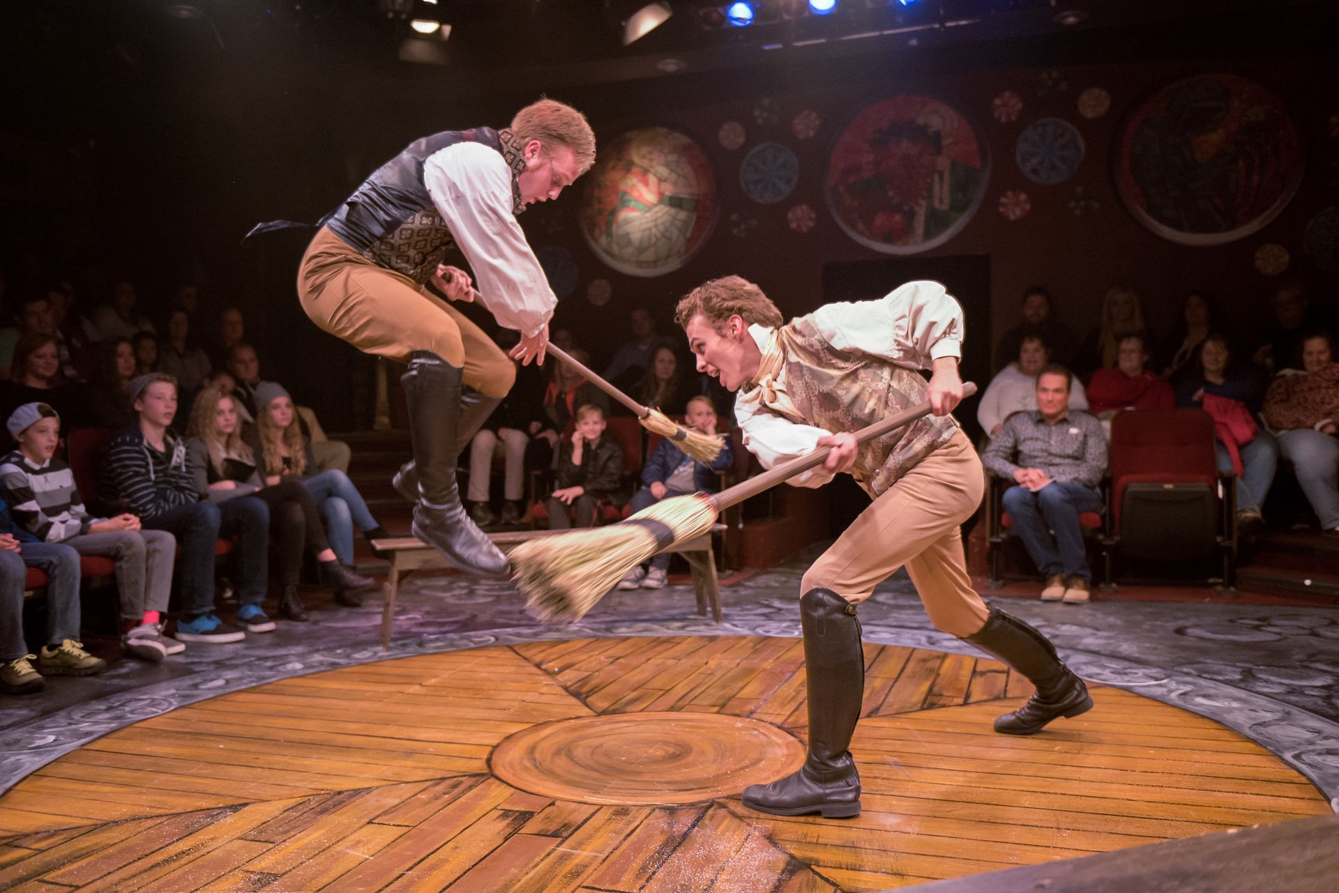 Two actors in period costumes fight with brooms on a circular wooden stage before a seated theater audience.