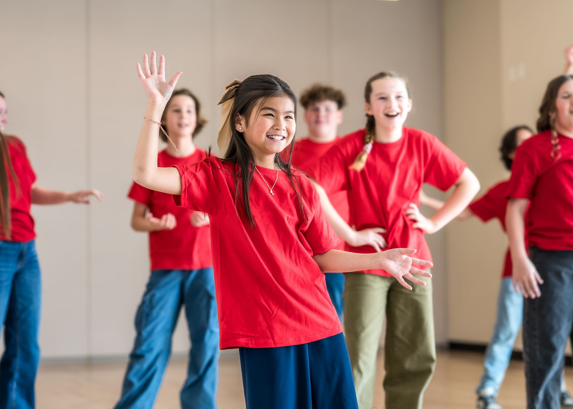A group of students in red shirts dancing and smiling in a studio, with one person in the foreground waving their hand.