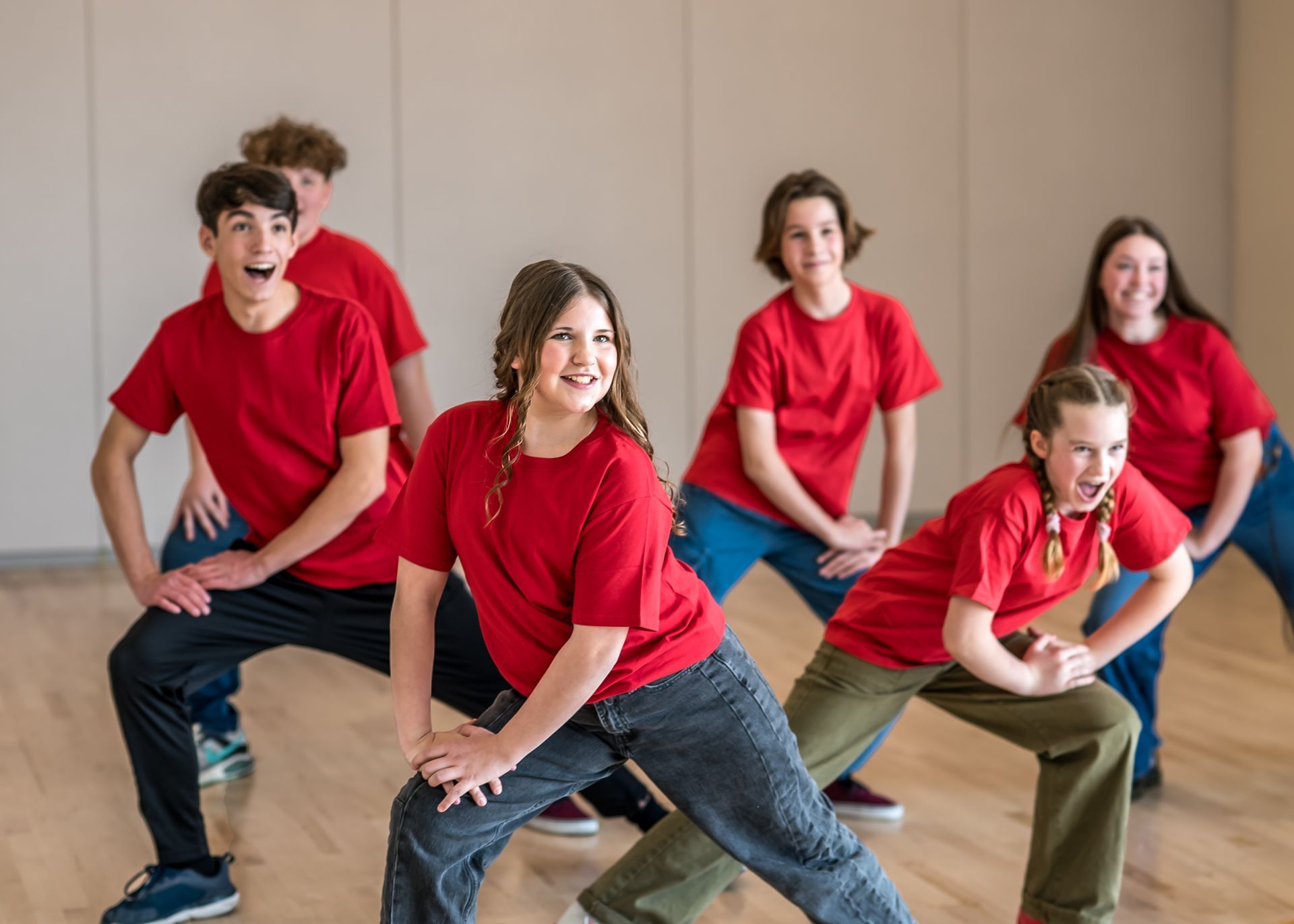 Six students wearing red shirts pose in a dance studio, performing a lunge with expressive faces.