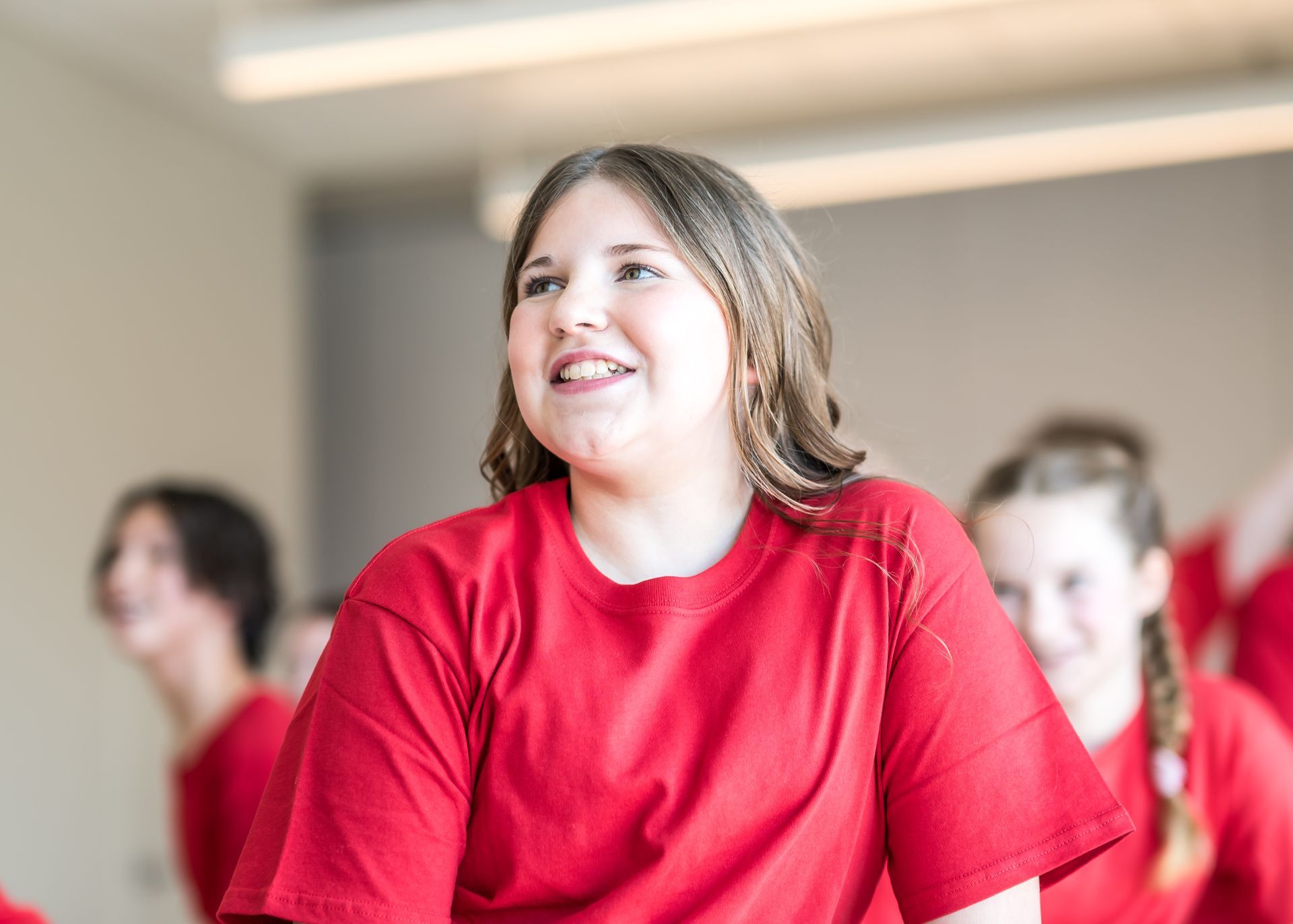 A person with long brown hair smiles while wearing a red t-shirt, with others in the background of a brightly lit room.